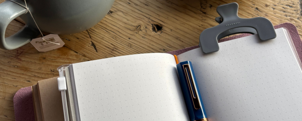 A journal is clipped open on a fresh page, on a wooden surface, a fountain pen laid upon it. A fresh pot of tea just visible to the top left.